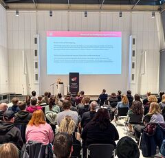 Großer Vortrag in einem modernen Veranstaltungssaal: Eine Person steht am Rednerpult vor einer Präsentationsfolie über Kinderrechte und Beteiligung, während ein großes Publikum auf Stühlen aufmerksam zuhört.